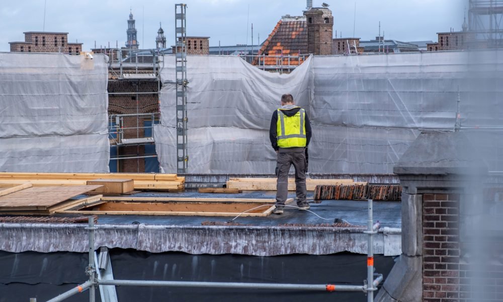 Building roof construction insulation works. Worker with yellow vest at rooftop, metal scaffolding. Amsterdam, Netherlands